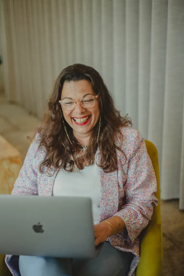 Mulher sorrindo usando óculos, com cabelo castanho e ondulado, sentada em uma cadeira amarela, usando uma blusa de lã...