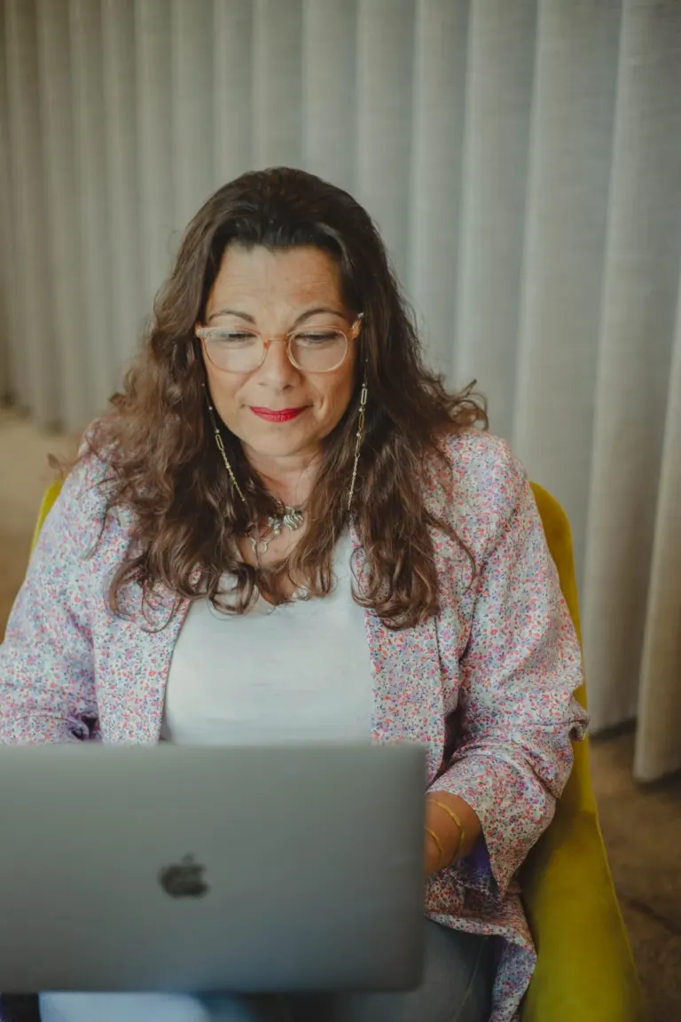 Mulher com cabelos cacheados, usando óculos e brincos longos, sentada em uma cadeira amarela, olhando para um laptop